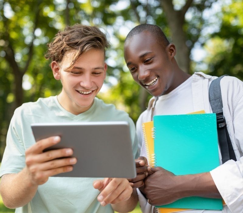 Student guy showing his project on digital tablet to friend, walking outdoors in college campus, looking at device and smiling. University lifestyle concept